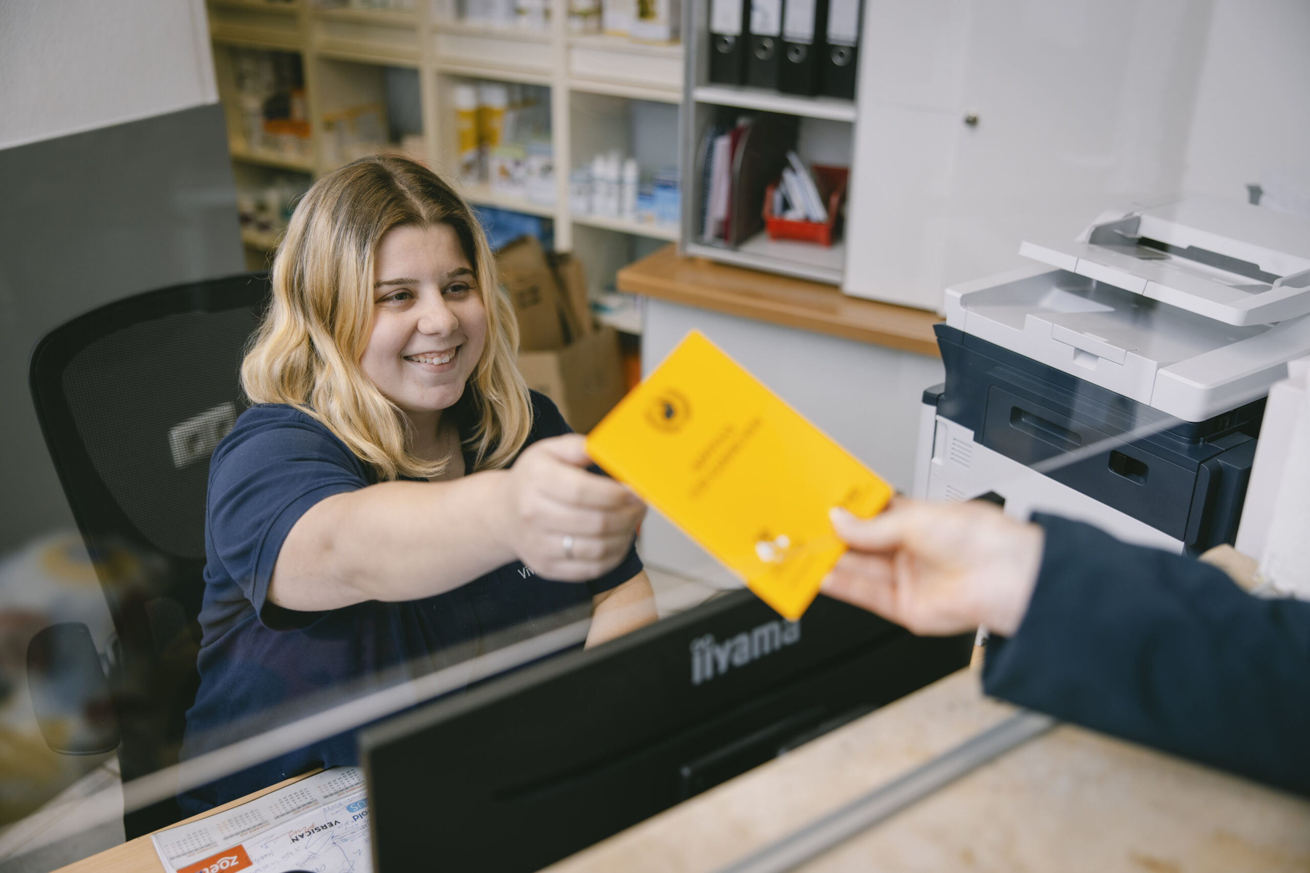 Eine Mitarbeiterin sitzt hinter dem Empfang und händigt einem Kunden den Impfpass des Tieres aus.